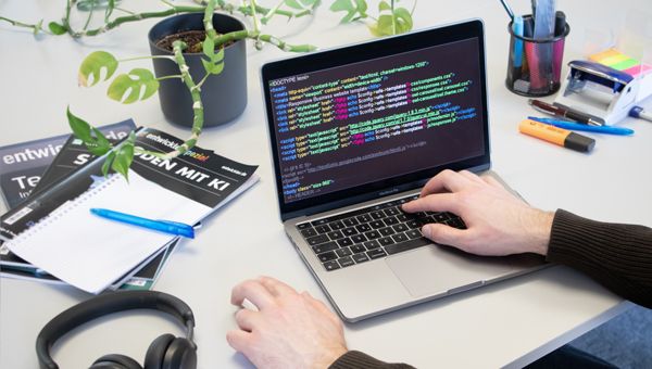 Person programmiert am Laptop mit Code, Notizbuch und Kopfhörern
